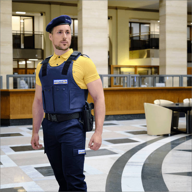 Uomo in uniforme di sicurezza, con gilet e berretto blu, in un ambiente interno.