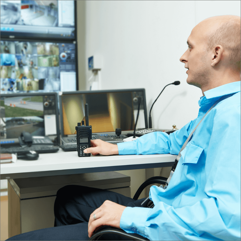 Uomo calvo in camicia azzurra, seduto davanti a più monitor di sorveglianza.